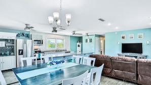 A modern kitchen with white cabinets, a dining area with a wooden table and white chairs, a brown leather sofa, and a flat-screen TV. - Sunny Upper Level 1 Block From 2 Beaches W/private Pool, High-speed Wi-fi (St. Augustine)