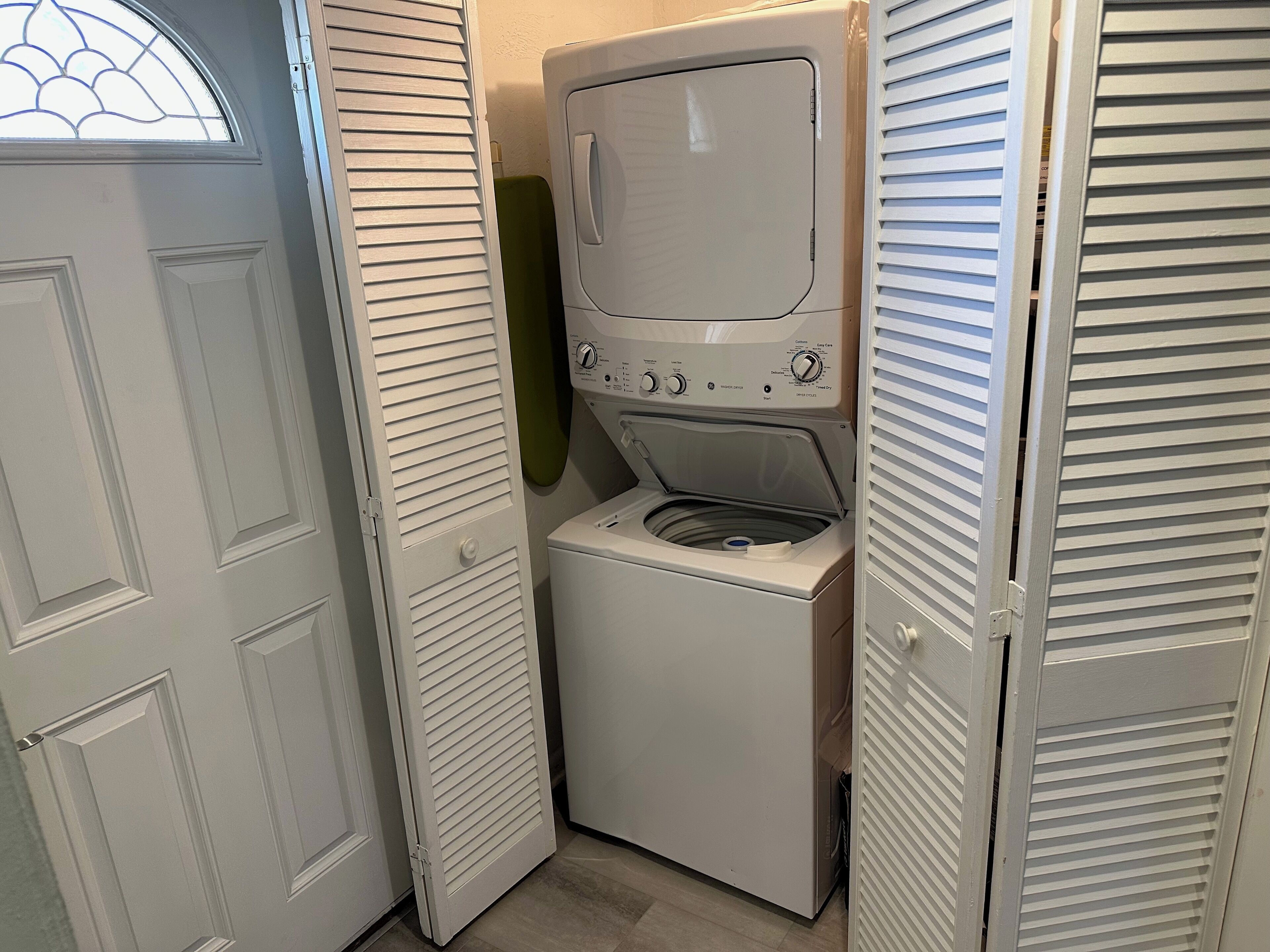 A compact laundry room with a front-loading washer and dryer, a door with a window, and a door with a louvered design.