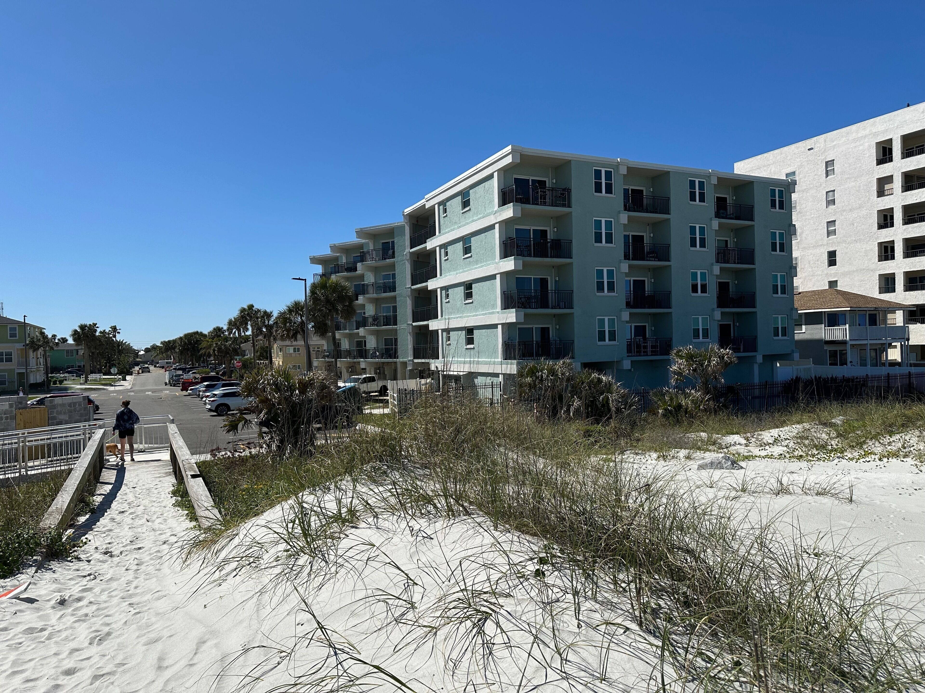 A beachfront building with multiple balconies, a sandy path leading to the beach, and a clear blue sky.