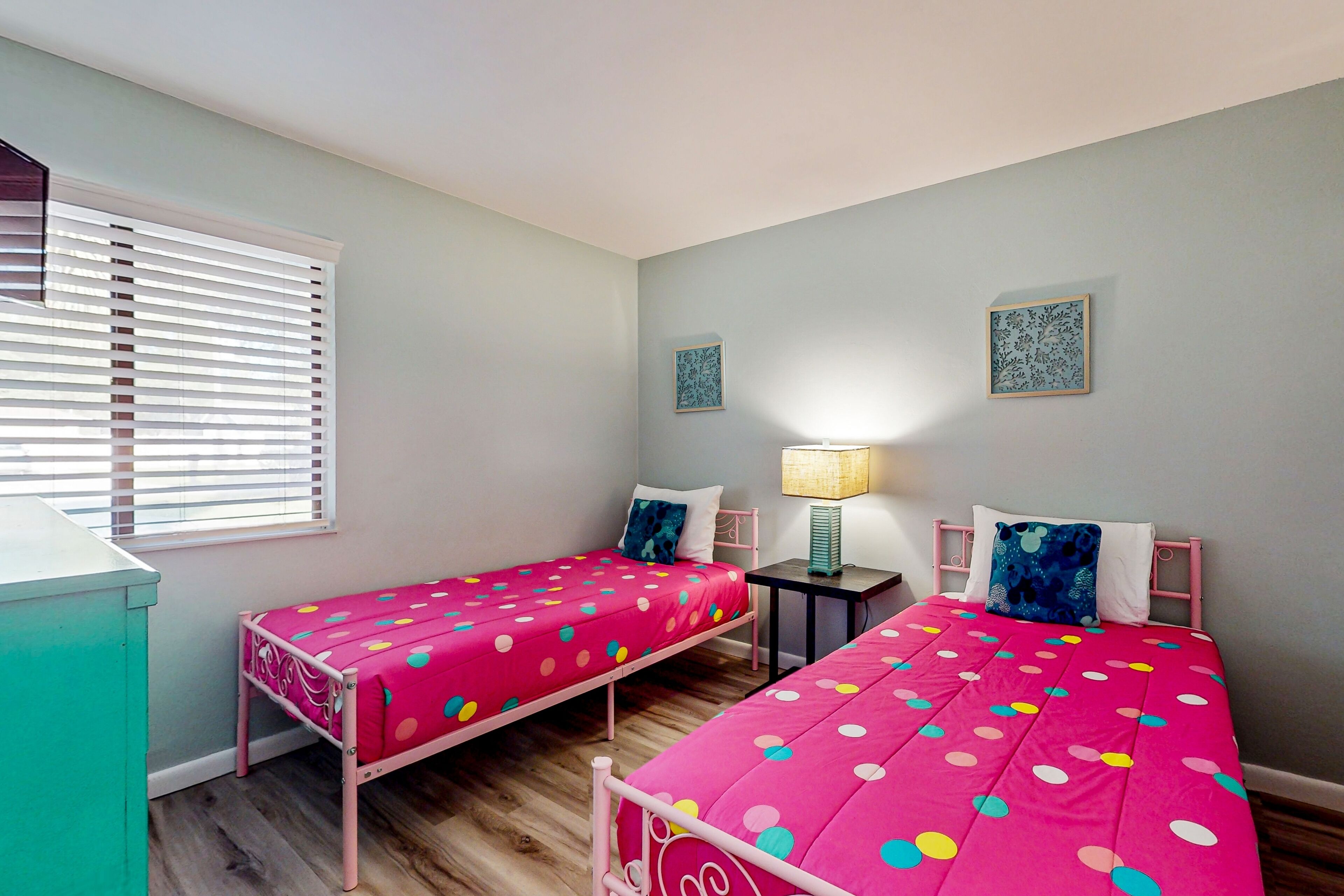 Un dormitorio con dos camas, una ventana con persianas, una mesita de noche con lámpara y dos cuadros colgados en la pared.