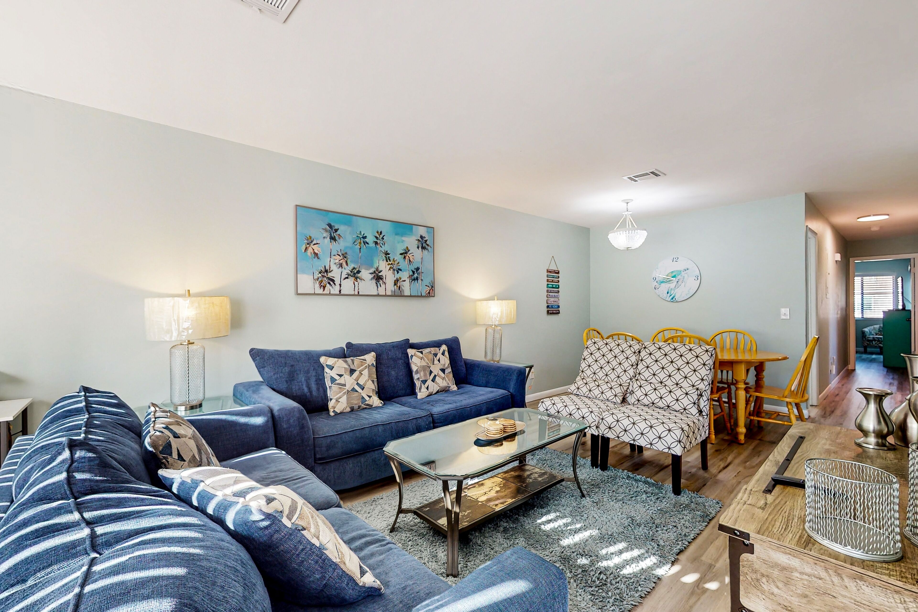 A living room with a blue sofa set, a glass coffee table, and a dining area with a yellow table and chairs.