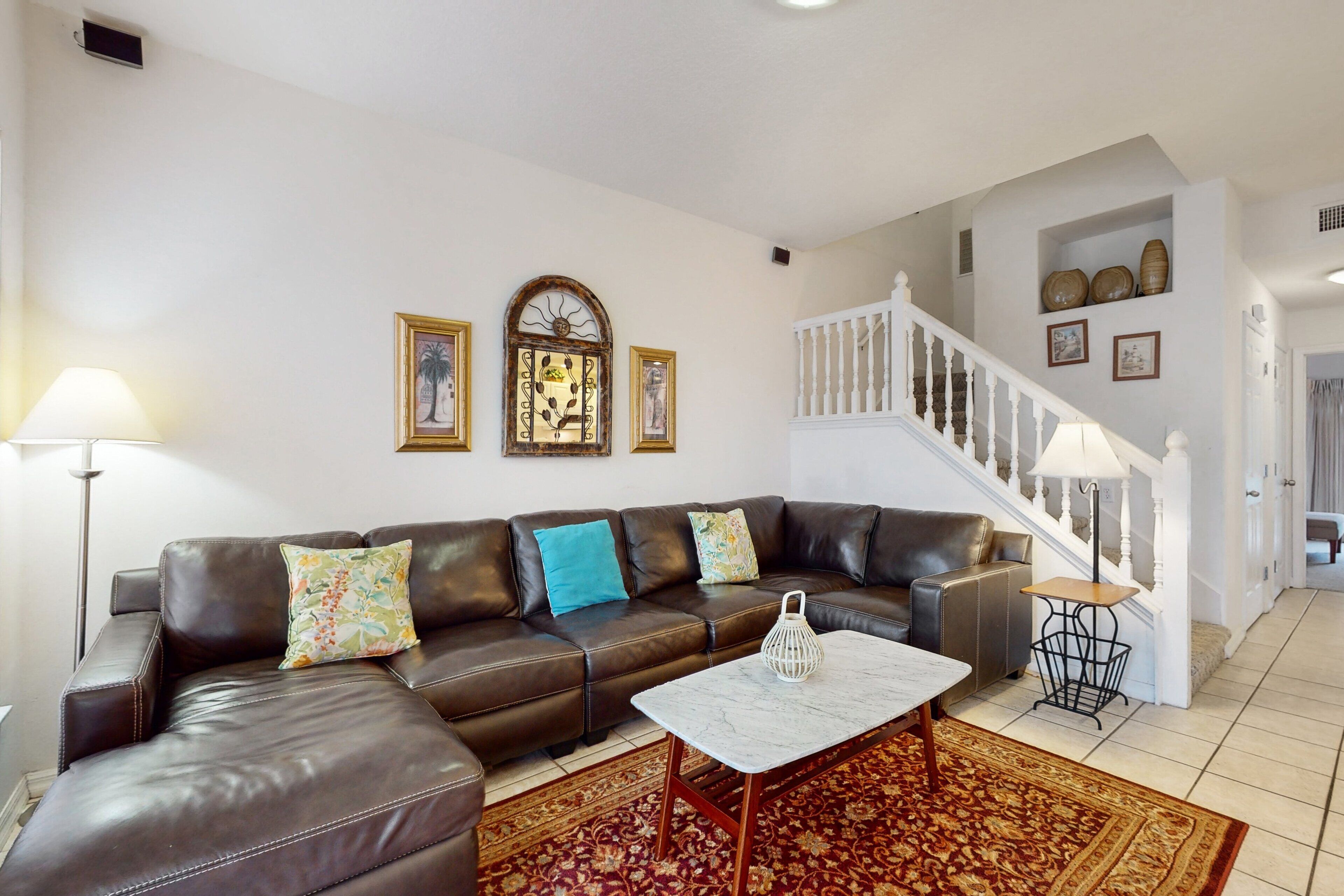 Ein Wohnzimmer mit braunem Ledersofa, einem weißen Couchtisch und einem gemusterten Teppich. Eine Treppe mit weißer Geländer führt zu einer weiteren Ebene.