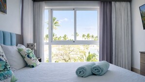 Ein Hotelzimmer mit großem Fenster, einem Bett mit weißer Bettwäsche und Blick auf Palmen.