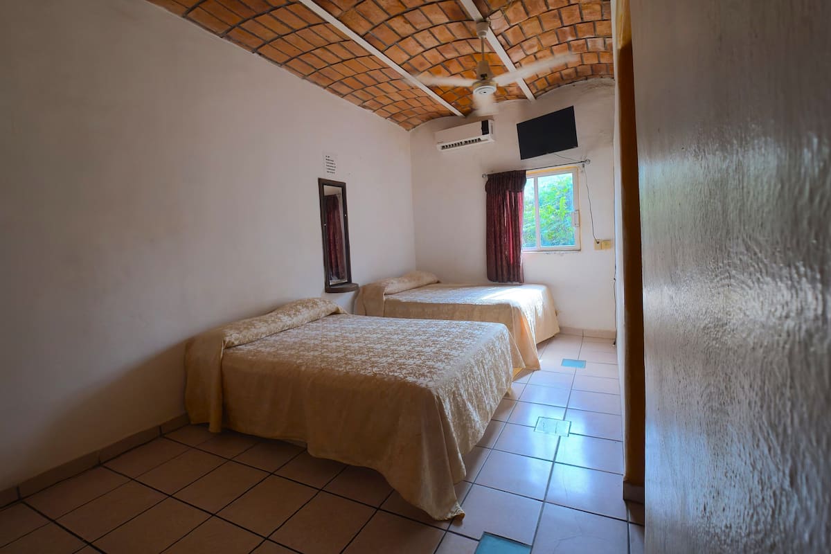 A room with two beds, a ceiling fan, an air conditioning unit, a window with curtains, and a wall-mounted TV.