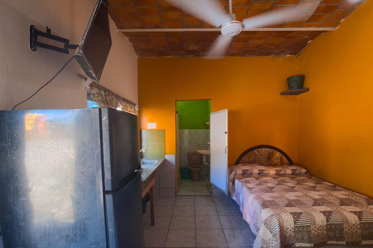 A compact room with a bed, a small desk, a refrigerator, and a ceiling fan.