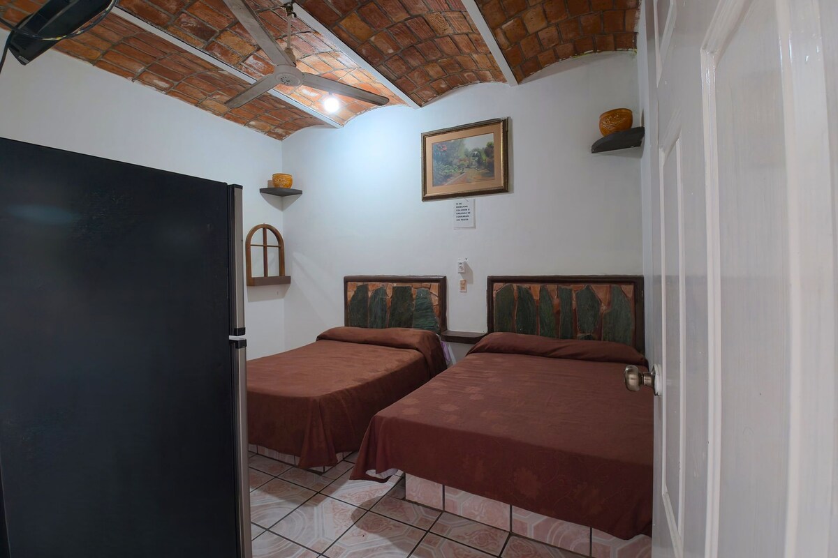 A room with two beds, a ceiling fan, a painting, and a brick ceiling.