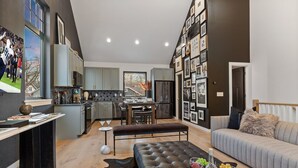 Une cuisine moderne avec des meubles de couleur grise, un coin repas avec une table et des chaises, et un salon avec un canapé et un pouf en cuir noir.