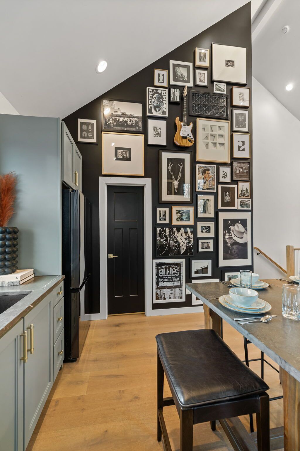 A modern kitchen with a black and white photo collage on the wall, a black refrigerator, and a wooden dining table with blue plates.