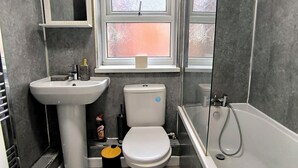 A bathroom with a white sink, toilet, and bathtub. There is a window above the toilet and a small shelf with a bottle.