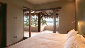 A bedroom with a large bed, a view of the beach, and a thatched roof structure.