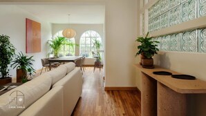 Een ruime woonkamer met een bank, eettafel en stoelen, een houten vloer en grote ramen met decoratieve glaspanelen.
