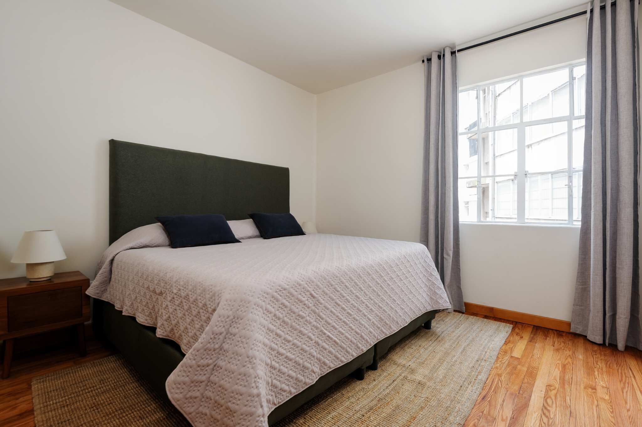 Une chambre à coucher avec un lit, une table de chevet, une fenêtre avec des rideaux et une vue sur les bâtiments à l’extérieur.