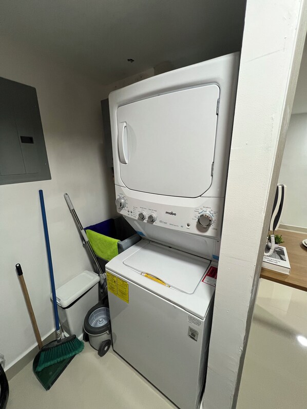 A compact laundry area with a washing machine and dryer, a broom, a dustpan, and a trash bin. - Luxury City View Apartments at Worldwide (Panama City)