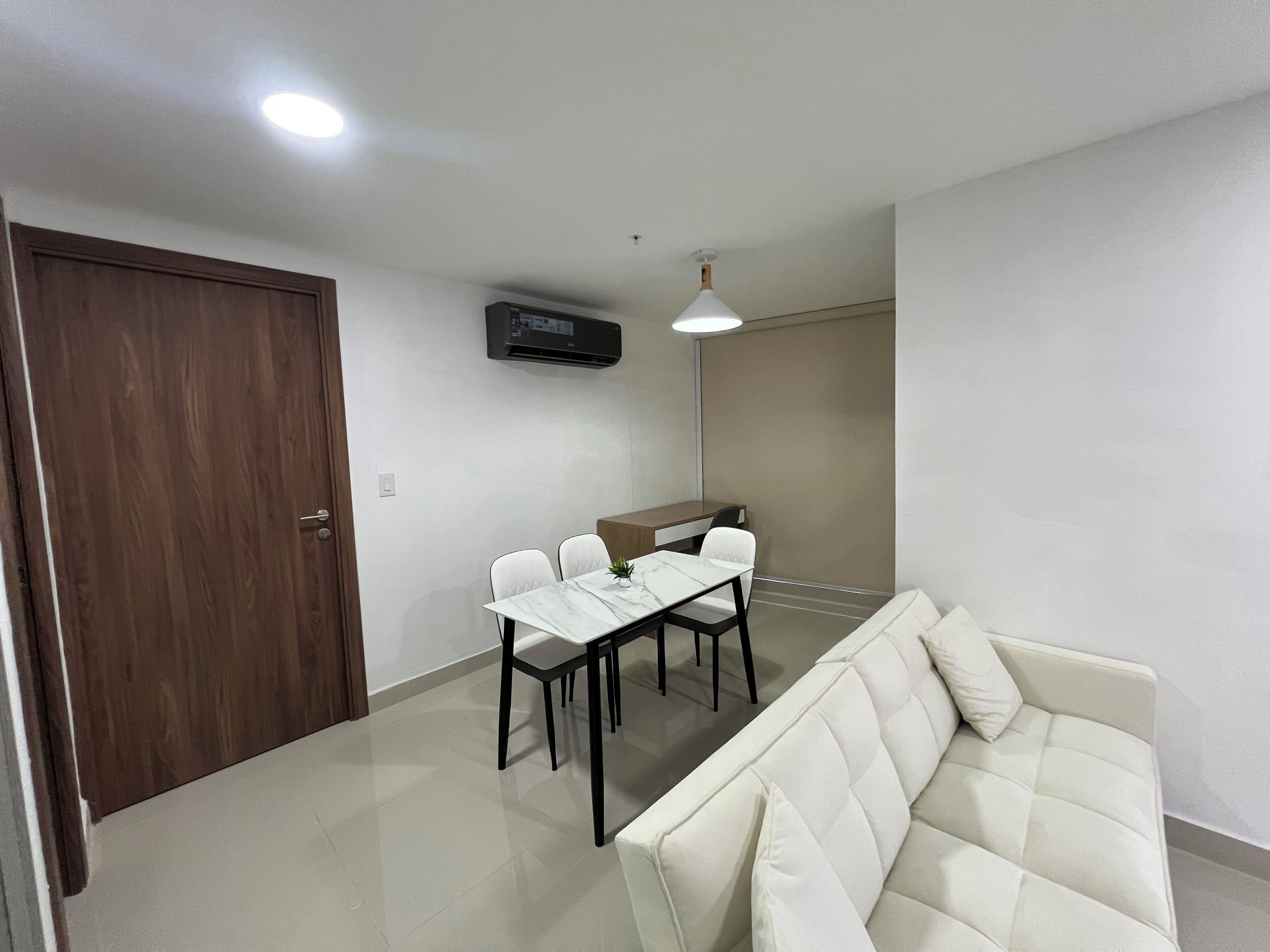 A modern living room with a white sofa, a dining table with chairs, and a small kitchen area with a refrigerator and a microwave.