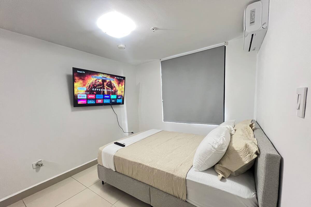 A bedroom with a bed, a TV mounted on the wall, a window with a blind, and a thermostat on the wall.
