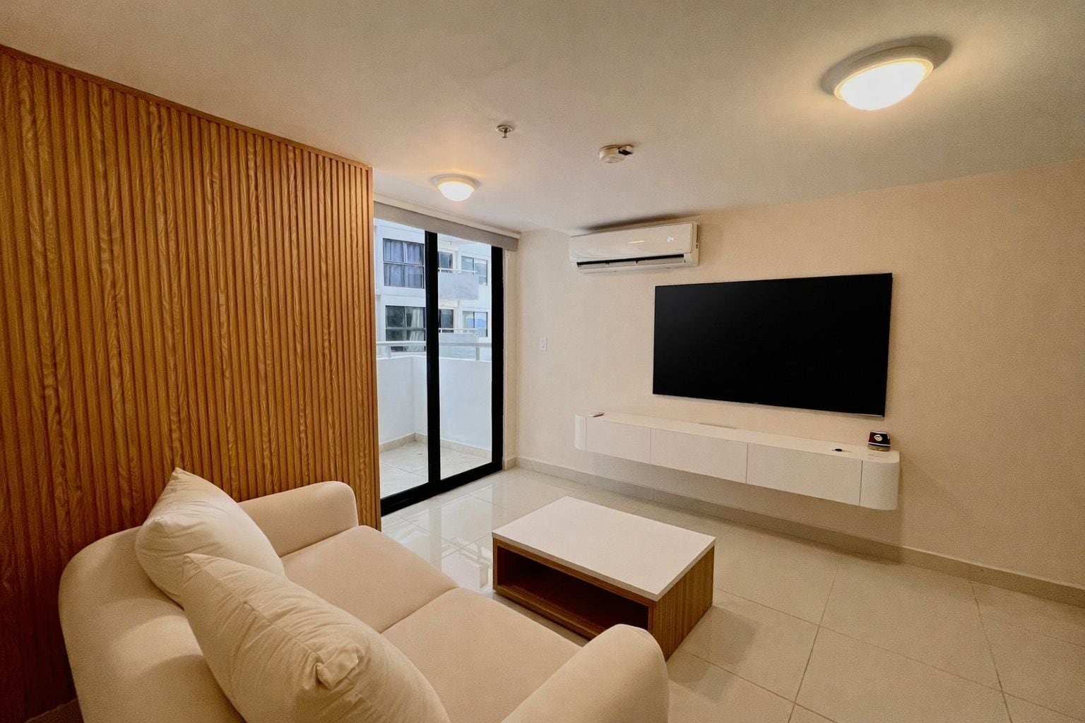 A modern living room with a sofa, a coffee table, a TV mounted on the wall, and an air conditioning unit.