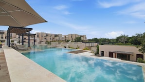 A swimming pool with lounge chairs, a covered seating area, and modern buildings in the background. - Gran Tulum Residences by Casago (Tulum)