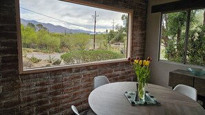 A dining area with a brick wall, a large window with a view of a desert landscape, and a table with a vase of tulips. - Quintessential Tucson - 2BR/2BA Entire House - Lower Foothills (Tucson)