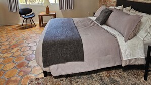 A bedroom with a bed, a chair, a table, a window with curtains, and a brick wall. - Quintessential Tucson - 2BR/2BA Entire House - Lower Foothills (Tucson)