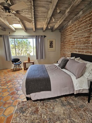 Ein Schlafzimmer mit Bett, Stuhl, Tisch, Fenster mit Vorhängen und einer Backsteinwand.
