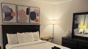 Ein ordentlich bezogenes Bett mit weißen Leinen, ein dunkles Kopfteil und ein Nachttisch mit einer Lampe. An der Wand über dem Bett hängen abstrakte Gemälde.