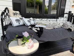 Ein Bett mit dunkler Decke, zwei Kissen mit Palmenmotiven, ein runder Tisch mit Tasse und Rosen sowie ein Fenster mit Jalousien.