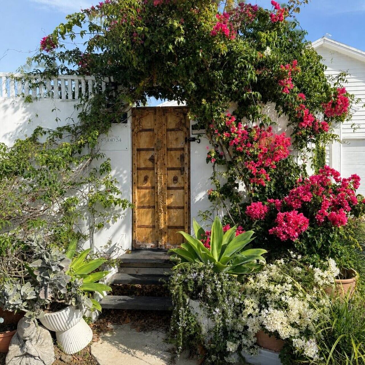 A white building with a wooden door, surrounded by lush greenery and flowering plants.
