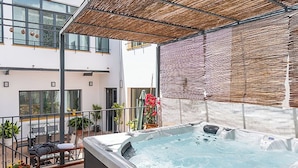 Ein Whirlpool auf einer Terrasse mit Tisch und Stühlen, umgeben von Topfpflanzen und einem Gebäude mit Balkonen.