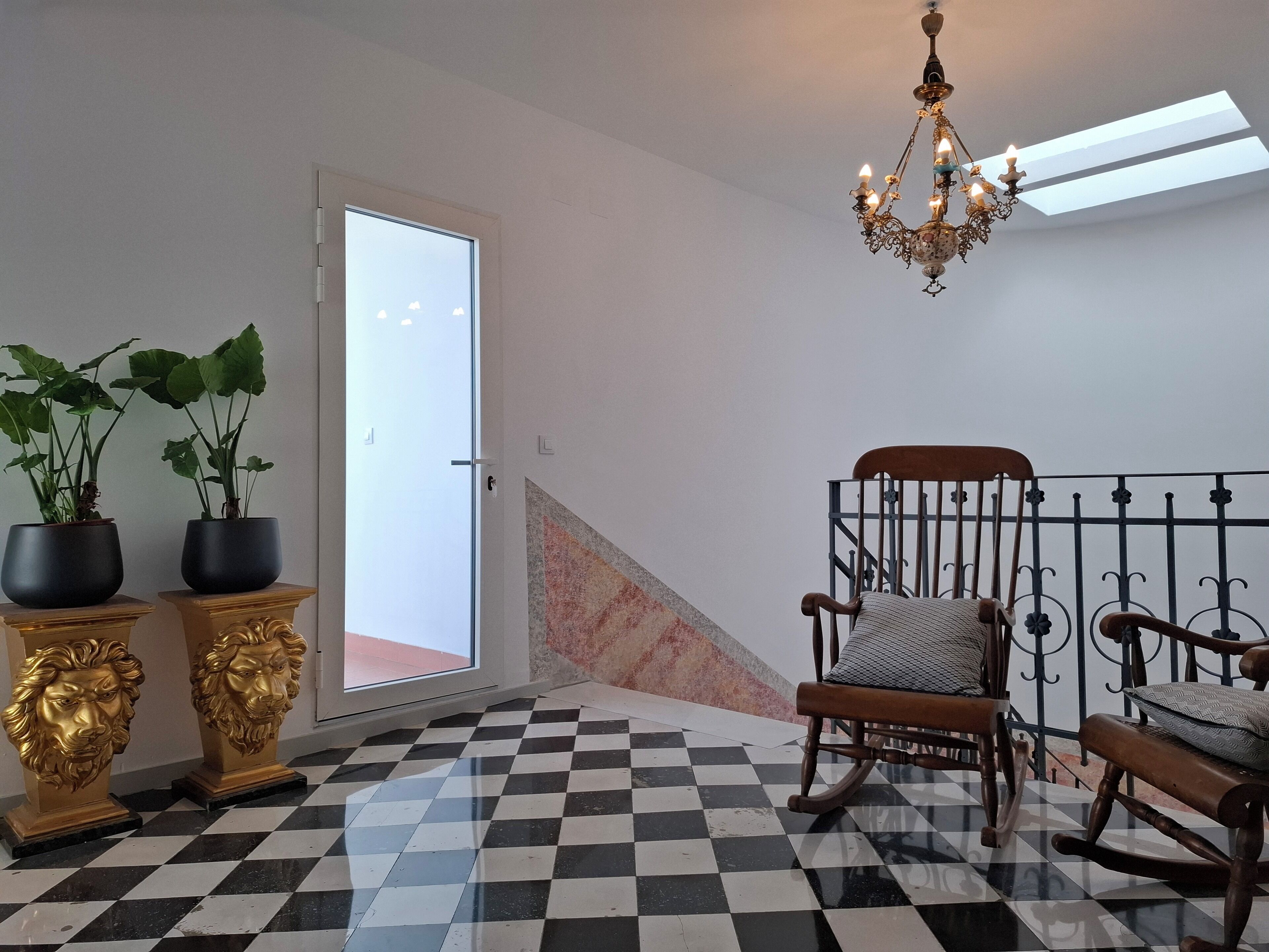 A room with a checkered floor, a chandelier, a rocking chair, and two potted plants on a pedestal.