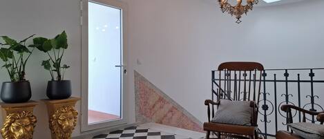 A room with a checkered floor, a chandelier, a rocking chair, and two potted plants on a pedestal.