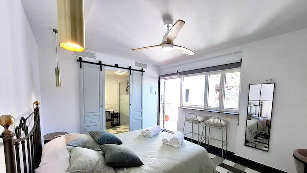 Ein Schlafzimmer mit Bett, Deckenventilator, Fenster und Spiegel. Ein Badezimmer ist durch eine offene Tür sichtbar.