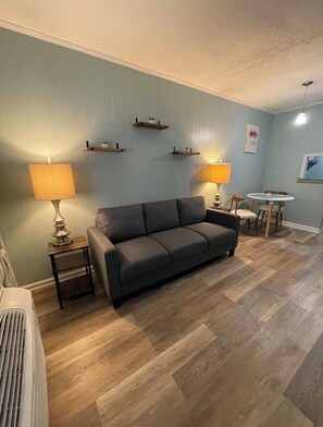 Ein Wohnzimmer mit grauem Sofa, zwei Beistelltischen mit Lampen, einem kleinen runden Tisch mit Stühlen und Holzböden.