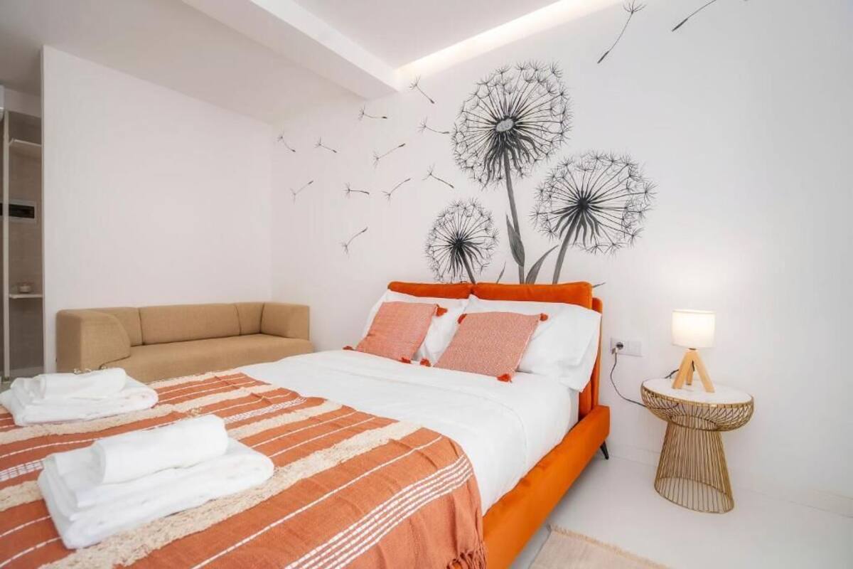 A modern bedroom with a bed, orange bedding, a white wall with dandelion artwork, a small round side table with a lamp, and a beige sofa.