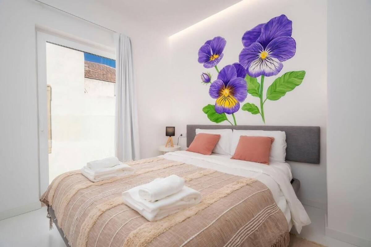 A hotel room with a large bed, white bedding, a grey headboard, and a wall mural of purple flowers.