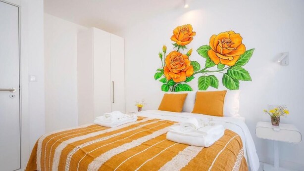 A hotel room with a bed, a nightstand, a white wardrobe, and a wall with a floral mural. - Primavera House - Sakura (Faro)