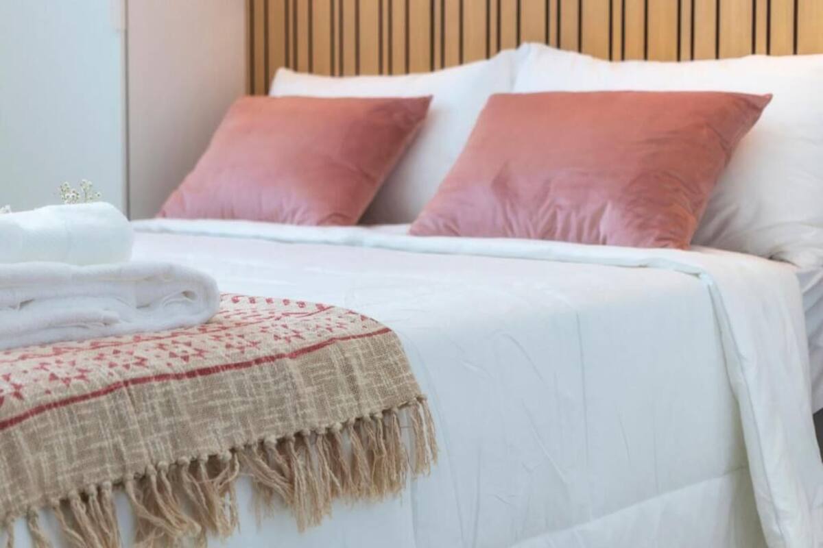 A neatly made bed with white and pink pillows, a beige throw blanket, and a folded white towel on the side.