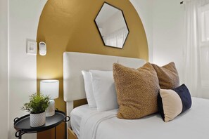 A neatly arranged hotel room with a white bed, multiple pillows, a small side table with a plant and lamp, and a wall-mounted mirror. - Stylish Retreat Near Mission Bay with Games & Firepit (San Diego)