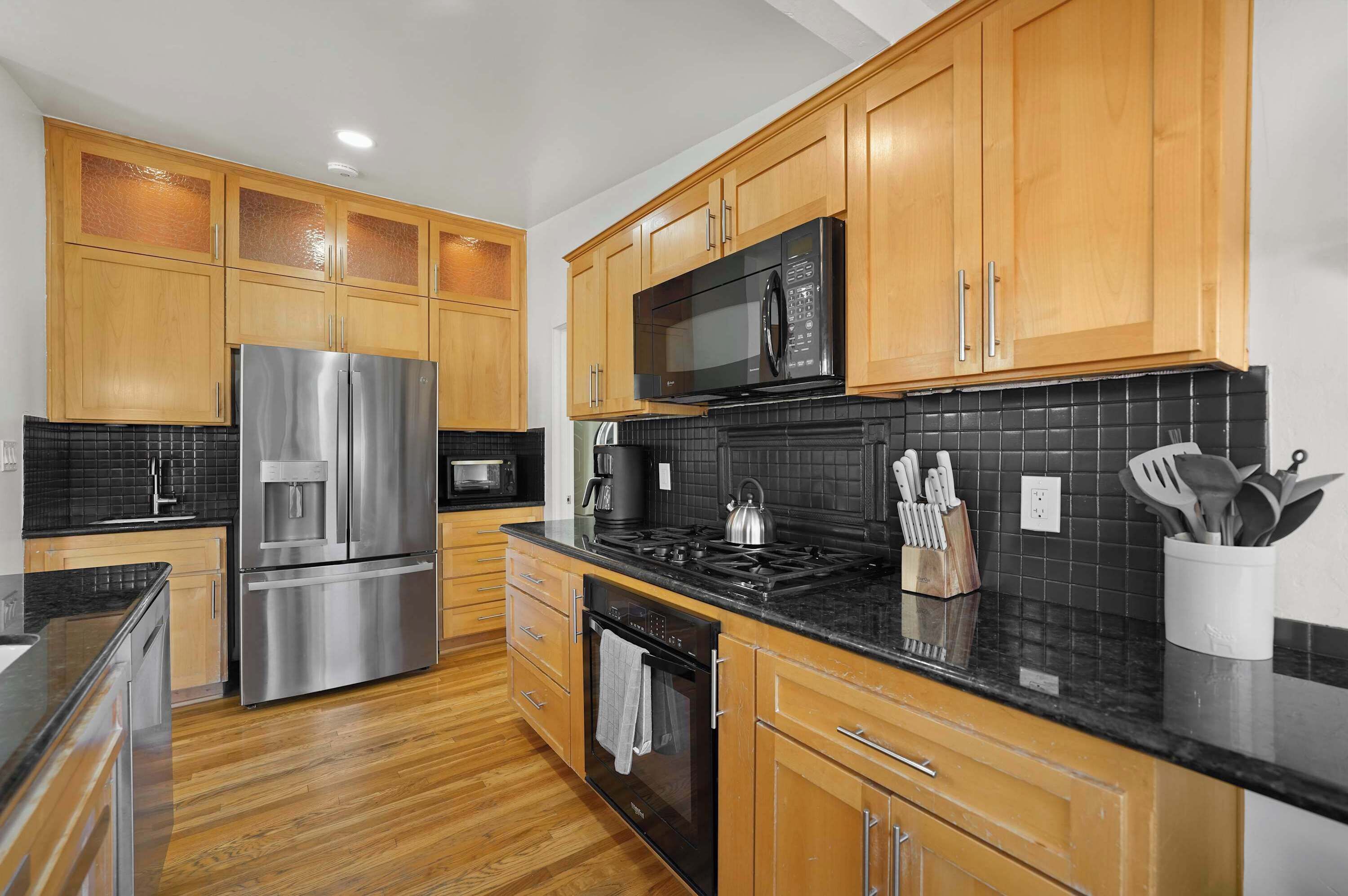 A modern kitchen with wooden cabinets, stainless steel appliances, a black tiled backsplash, and a wooden floor.
