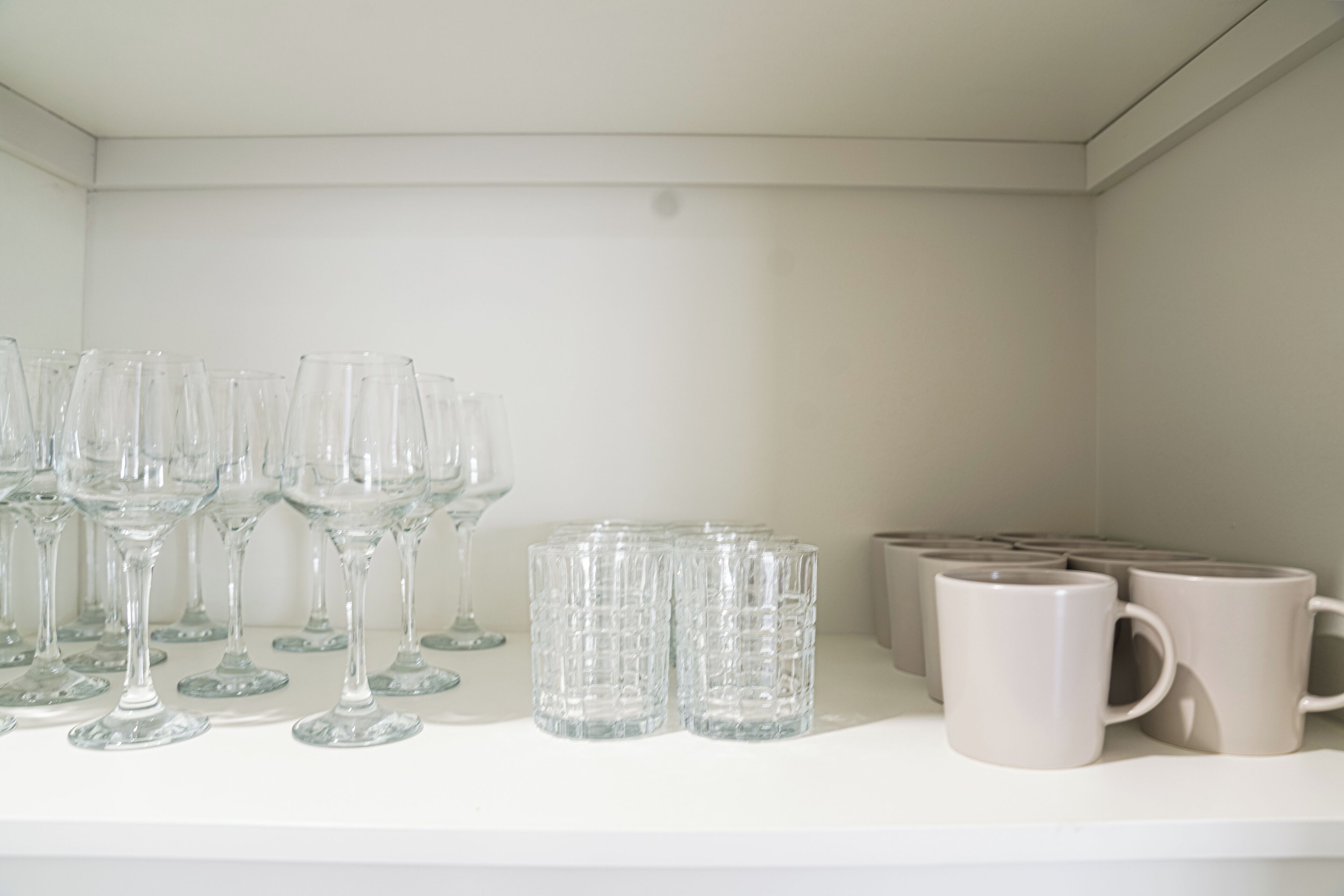 A white shelf with clear glassware and beige mugs.