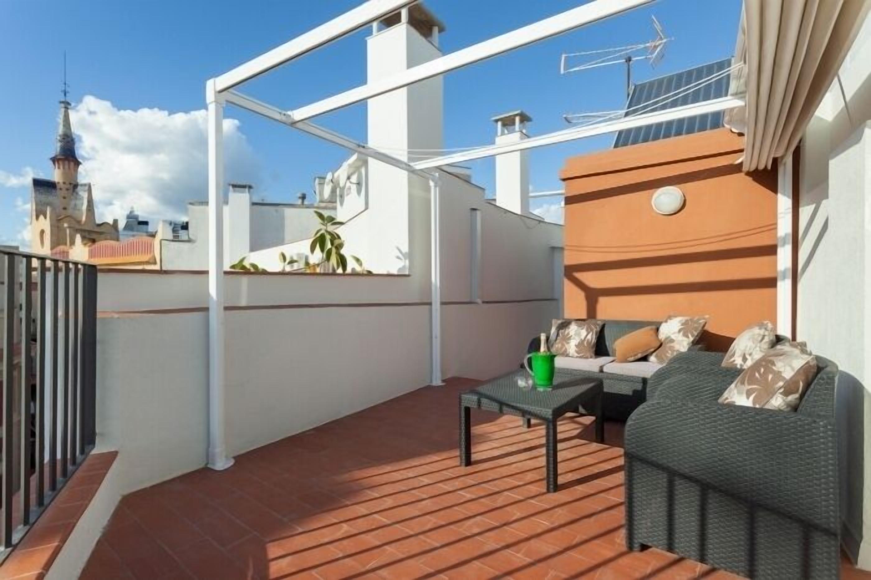 Eine Dachterrasse mit einer Couch, einem Tisch, einem grünen Eimer und Blick auf Stadtgebäude.