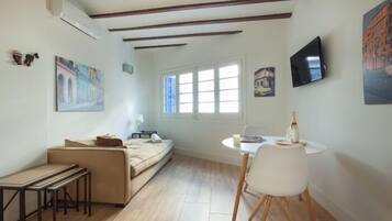 A room with a sofa, a dining table with chairs, a TV mounted on the wall, and a window providing natural light.