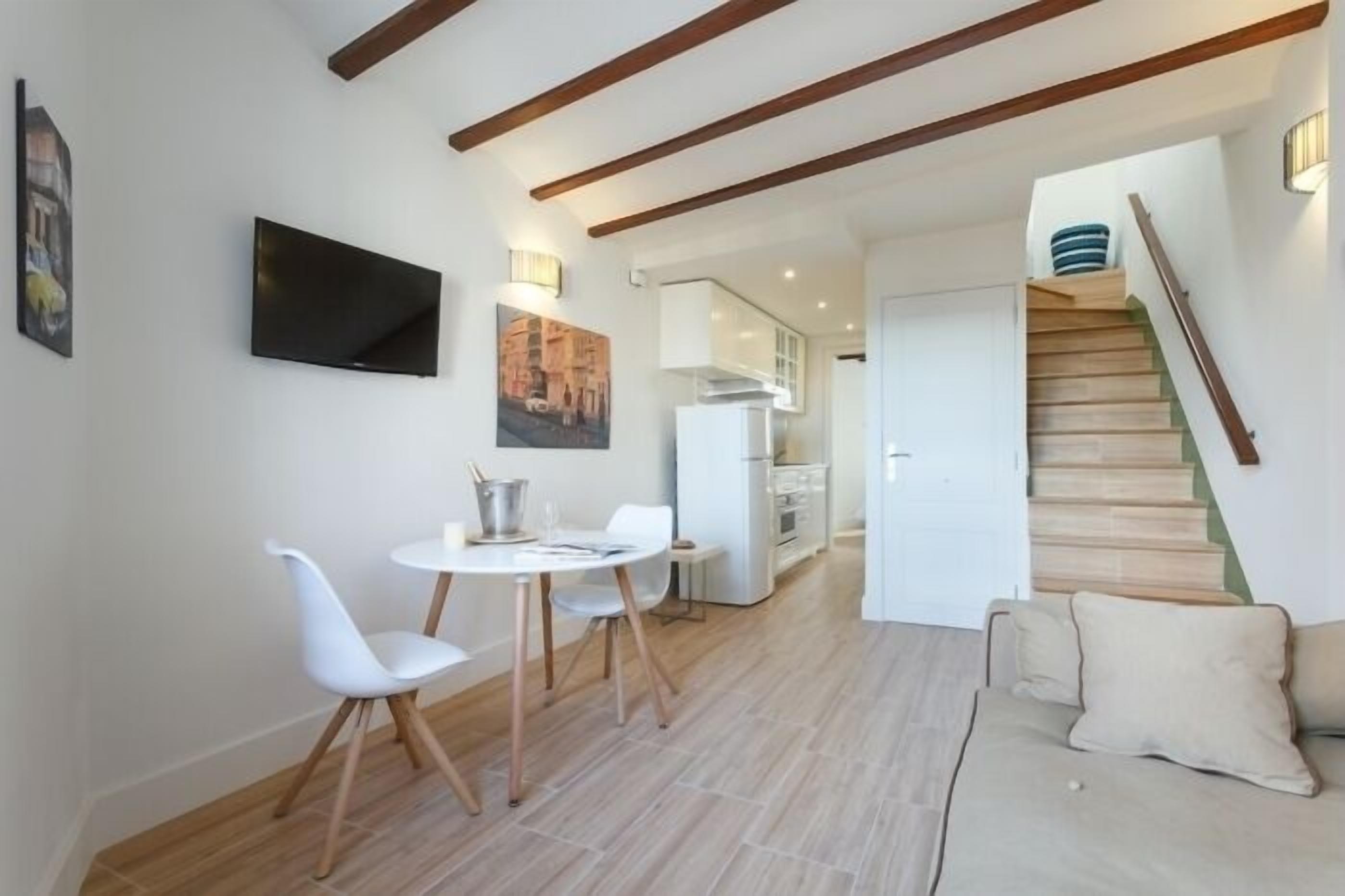 En moderne stue med sofa, spisebord med stole, et vægmonteret fladskærms-tv og en trappe, der fører til en etage ovenover.