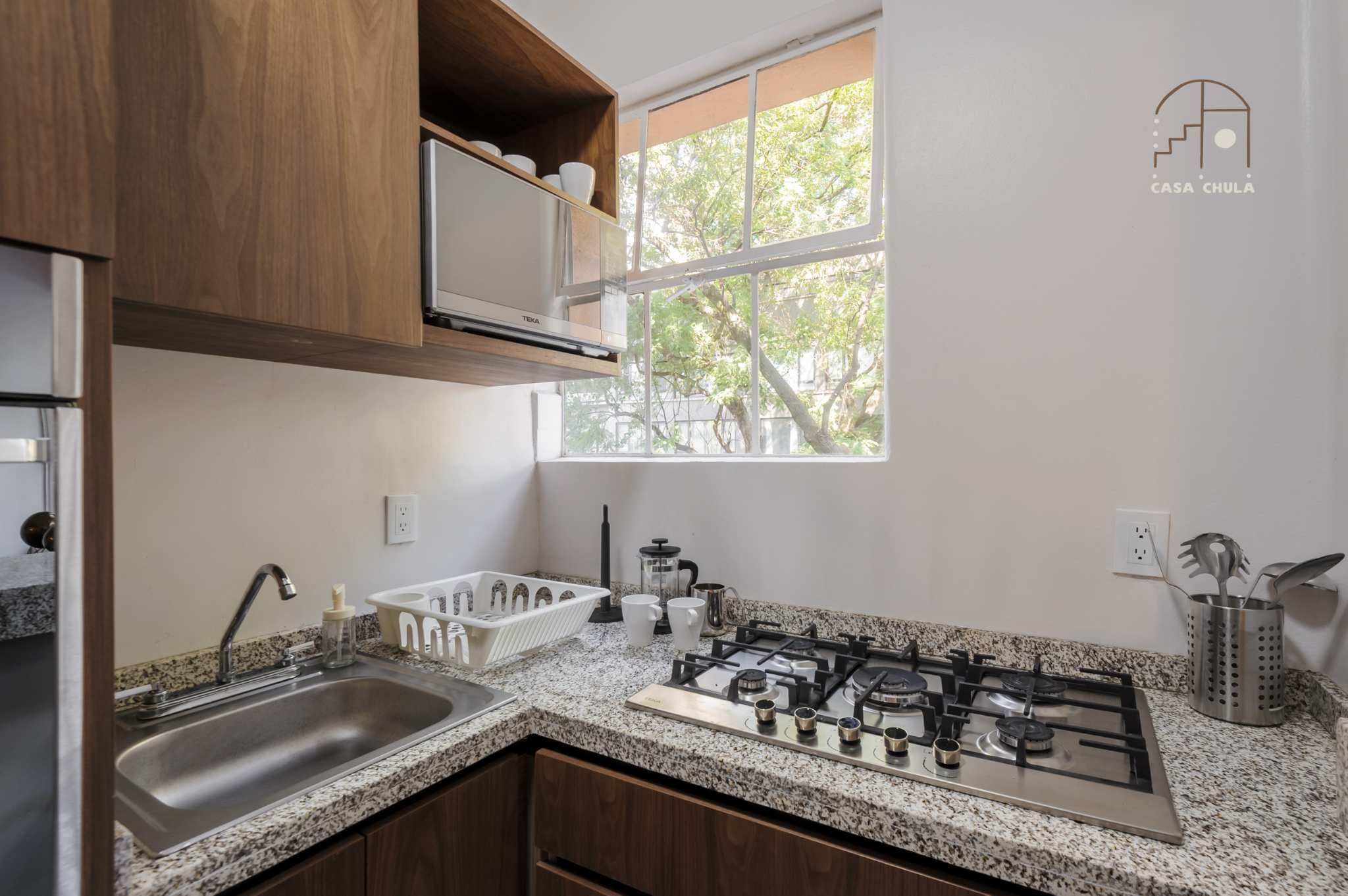 A kitchen with granite countertops, a stainless steel sink, a gas stove, wooden cabinets, and a window with a view of trees.