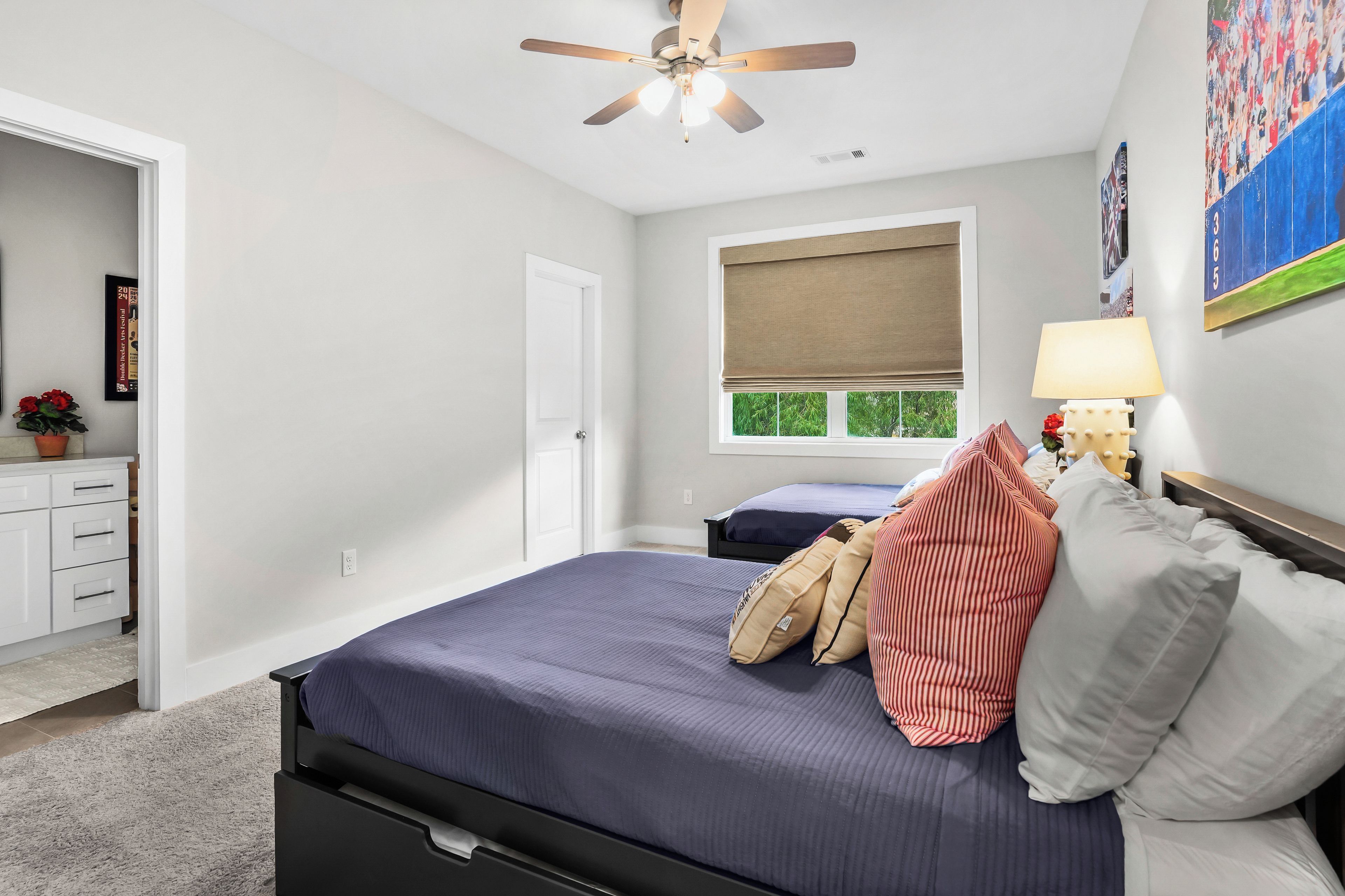 A bedroom with a bed, a ceiling fan, a window with a view, a bedside table with a lamp, and a dresser.