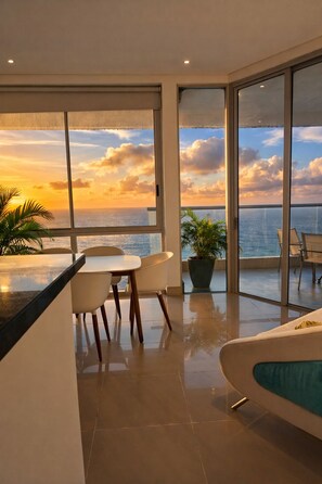 A modern living room with a large window overlooking the ocean at sunset, a dining area with chairs, and a potted plant. - 3BR Ocean Gem: Breakfast & Daily Cleaning Optional (Cartagena de Indias)