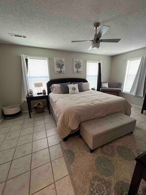 A bedroom with a bed, a ceiling fan, a bench, a nightstand, a lamp, and a window with curtains.
