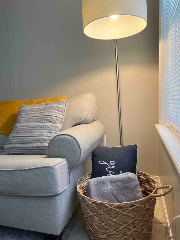 A corner of a room with a grey sofa, a floor lamp, a basket with a towel, and a window with blinds.