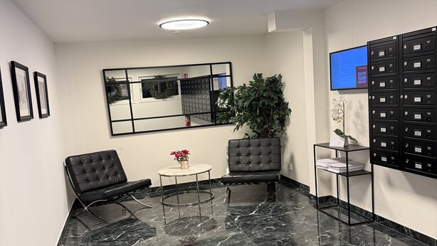 En moderne lobby med et marmorgulv, sorte læderstole, et lille bord med en vase, et stort spejl og et digitalt display på væggen.