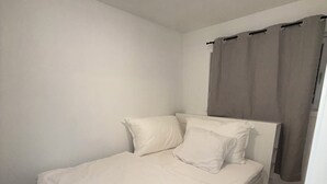 Et lille værelse med en enkelt seng, et hvidt sengetæppe, en loftlampe og et vindue med gardiner.
