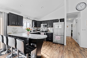 A modern kitchen with dark cabinets, a central island with bar stools, stainless steel appliances, and a clock on the wall.